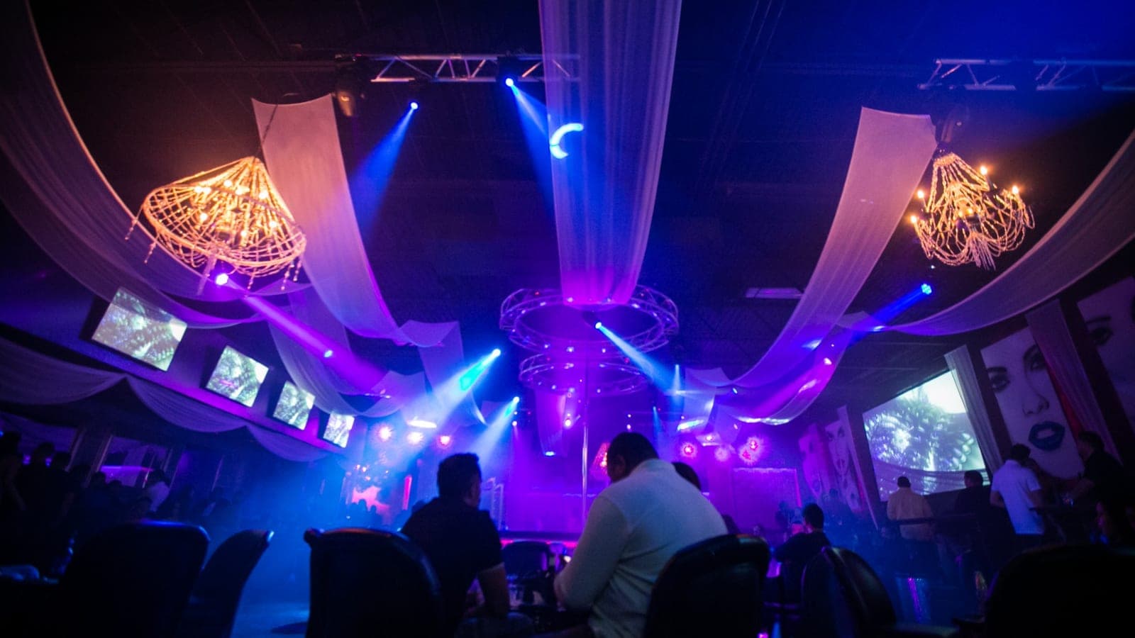 Two crystal chandeliers and white ceiling drapes over the main floor, stage lights cutting through haze above seated guests