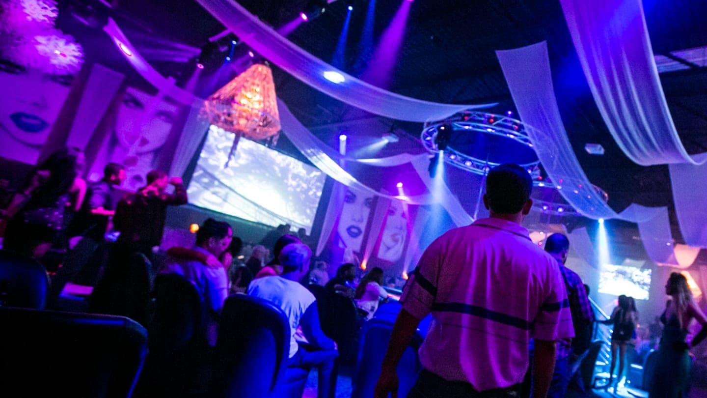 Guests on a busy main floor under purple light, with a chandelier and a video screen behind them
