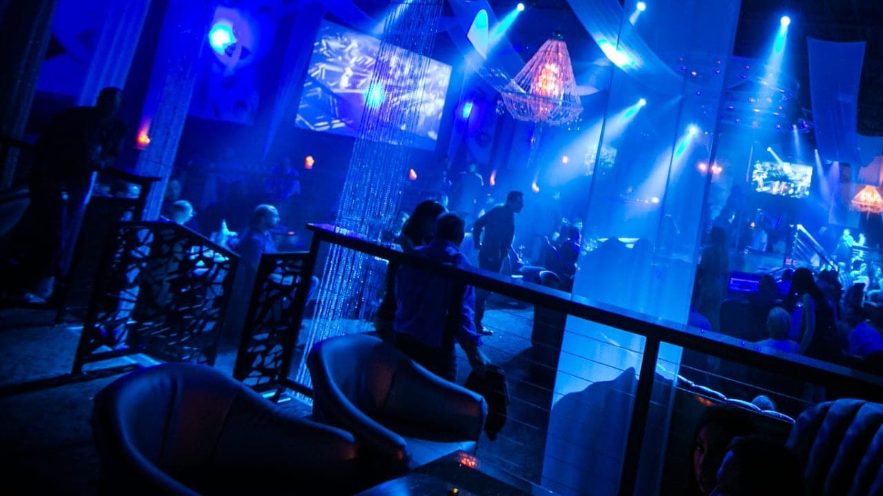 Armchairs and a railing in the blue-lit VIP lounge, a video screen glowing beyond